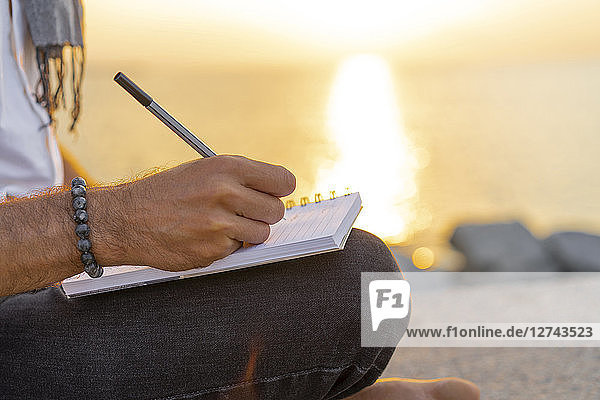Spain. Man writing on a notebook during sunrise on rocks at the beach
