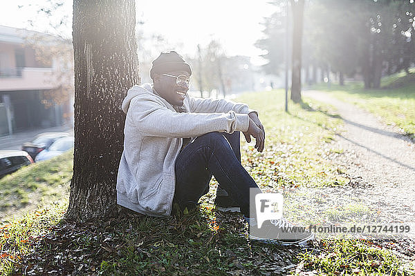 Laughing young man sitting in park  leaning on tree trunk