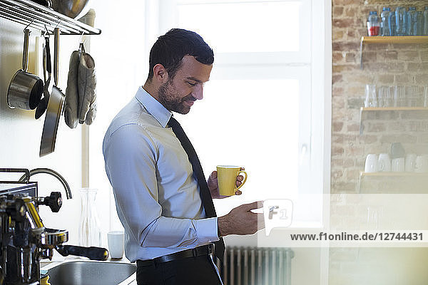 Businessman having coffee break in office kitchen looking at cell phone