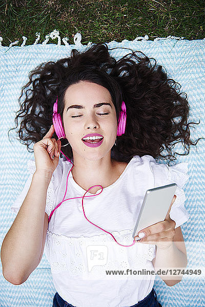 Portrait of young woman lying on blanket listening music with smartphone and pink headphones