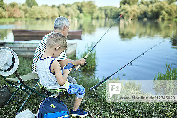 Little boy sitting with grandfather at lakeshore testing smartphone