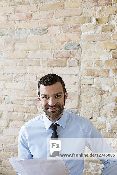 Portrait of smiling businessman with documents at brick wall