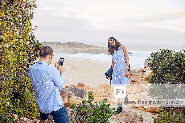Young man with smart phone photographing girlfriend with ocean in background