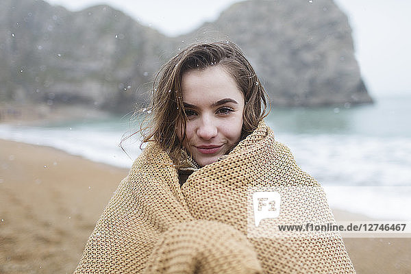 Portrait smiling teenage girl wrapped in blanket on snowy winter beach