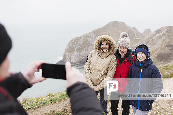 Mann mit Fotohandy fotografiert Familie in warmer Kleidung auf einer Klippe mit Blick auf das Meer