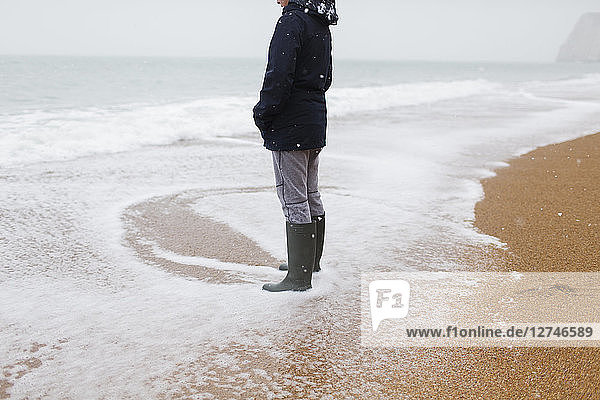 Teenage boy in rubber boots standing in snowy winter ocean surf