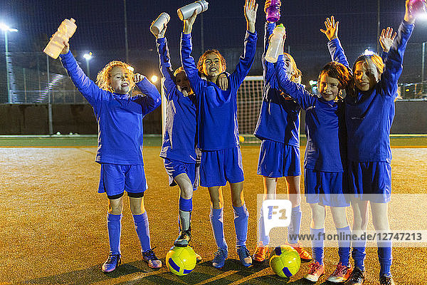 Porträt einer selbstbewussten Mädchenfußballmannschaft mit Wasserflaschen  die nachts auf dem Spielfeld jubeln