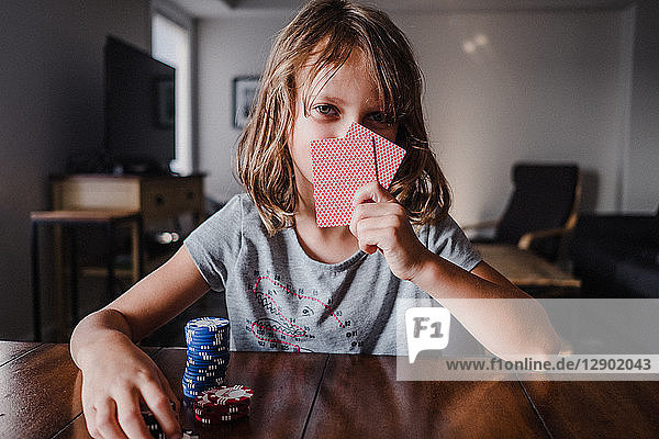Mädchen  das sich hinter Spielkarten mit Spielchips am Tisch versteckt  Portrait