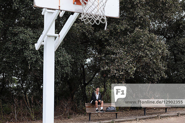 Männlicher jugendlicher Basketballspieler sitzt auf einer Parkbank am Basketballfeld