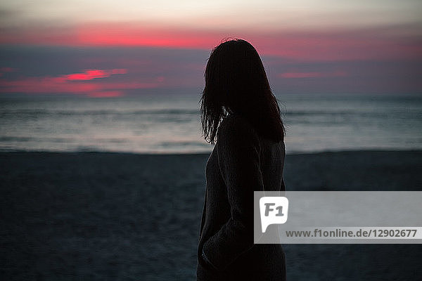 Junge Frau  die allein am Strand steht und den Sonnenaufgang beobachtet  Rückansicht