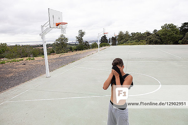 Male teenage basketball player wiping his brow with vest on basketball court