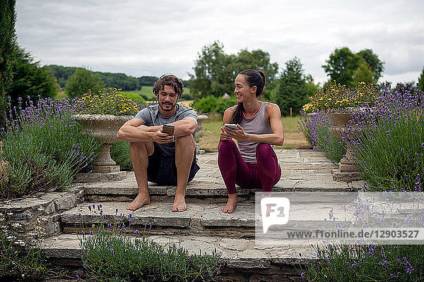 Mann und Frau üben Yoga im Garten und schauen sich auf der Terrasse Smartphones an