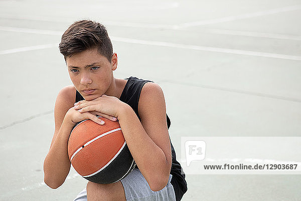 Männlicher jugendlicher Basketballspieler mit Ball auf dem Basketballfeld kauernd  Porträt