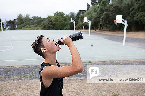 Männlicher jugendlicher Basketballspieler  der auf dem Basketballplatz aus einer Wasserflasche trinkt