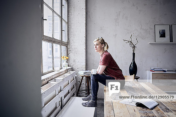 Smiling woman with coffee mug sitting on desk in loft looking through window