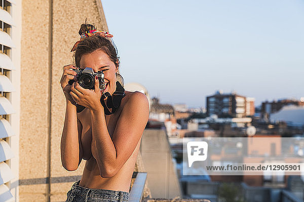 Happy topless young woman with camera taking pictures on balcony