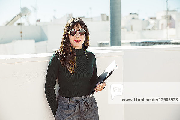 Businesswoman with sunglasses standing on rooftop  holding laptop