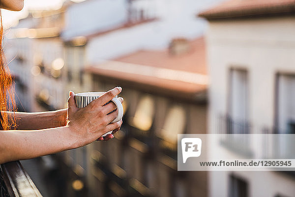 Hands of woman on balcony above the city at sunset holding cup of coffee