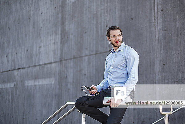 Businessman with smartphone and earbuds sitting on a railing