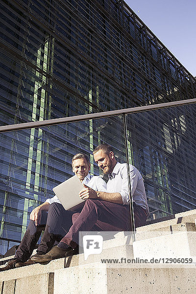 Two smiling businessmen sitting on stairs using laptop