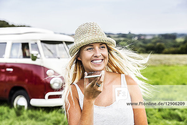 Happy young woman with cell phone in front of van in rural landscape