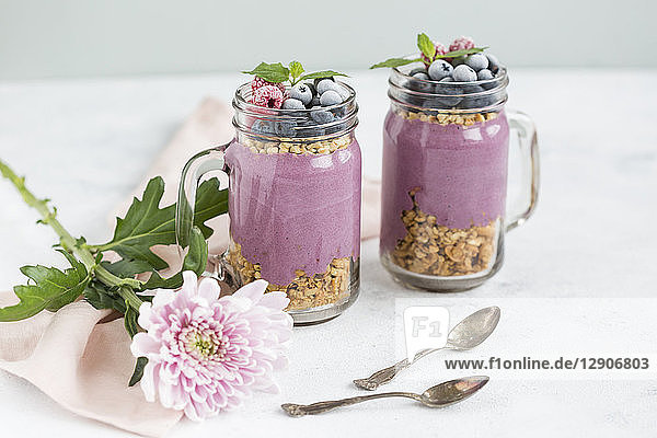 Two glasses of yoghurt with peanut granola  aronia powder and topping of chopped hazelnuts and frozen berries