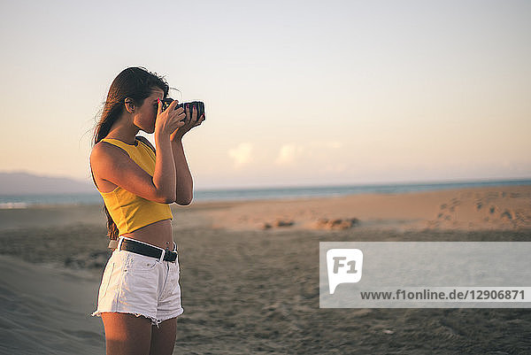 Teenage girl taking photos with camera on the beach at sunset