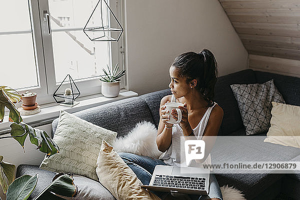 Young woman with laptop and cup of coffee sitting on the couch at home looking out of window