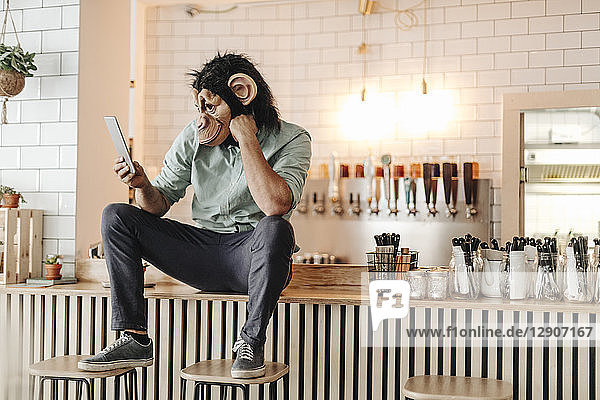 Man wearing monkey mask  sitting on counter of a bar  using digital tablet