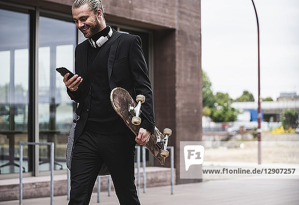 Smiling fashionable young man holding cell phone and skateboard passing office building