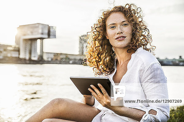 Germany  Cologne  portrait of young woman with tablet sitting at riverside