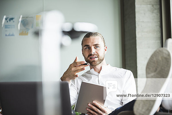 Young businessman sitting in office with feet up holding tablet and talking
