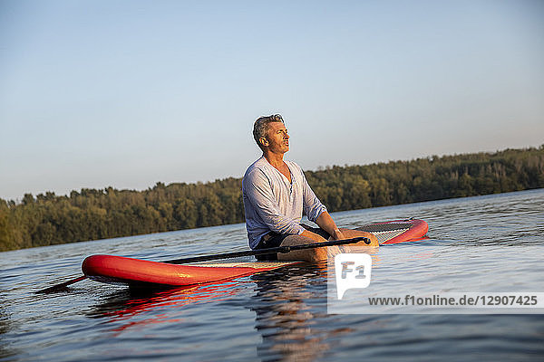 Mature man sitting on paddleboard on a lake by sunset relaxing