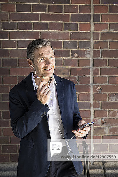 Mature businessman with earphones and smartphone at brick wall