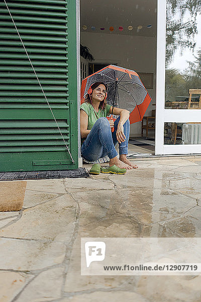Smiling mature woman sitting on terrace in rain under umbrella listening to music with headphones