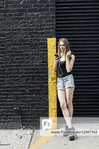 Young woman leaning on street corner  listening music  using smartphone