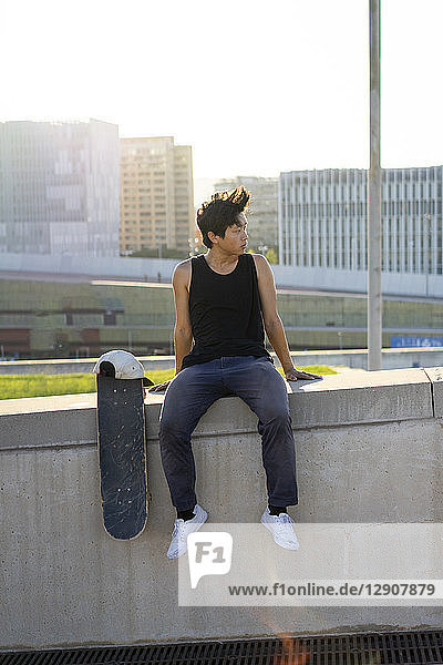 Young man sitting on urban wall next to skateboard at sunset