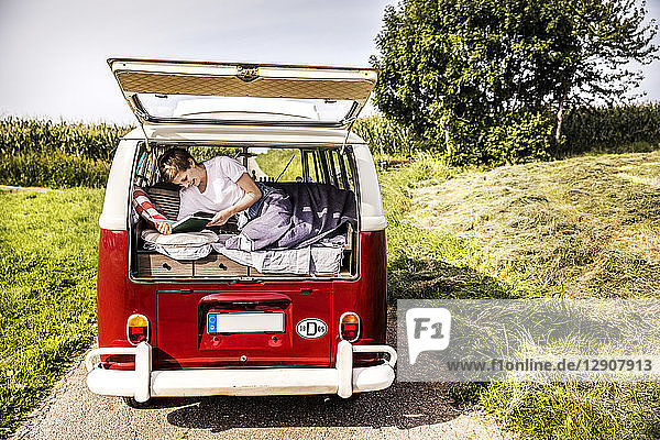 Happy woman lying in a van in rural landscape reading book