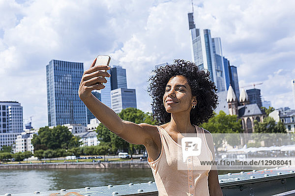 Germany  Frankfurt  portrait of content young woman with curly hair taking selfie in front of skyline