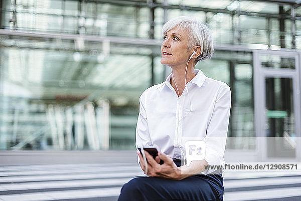Senior businesswoman sitting outside with smartphone and earbuds