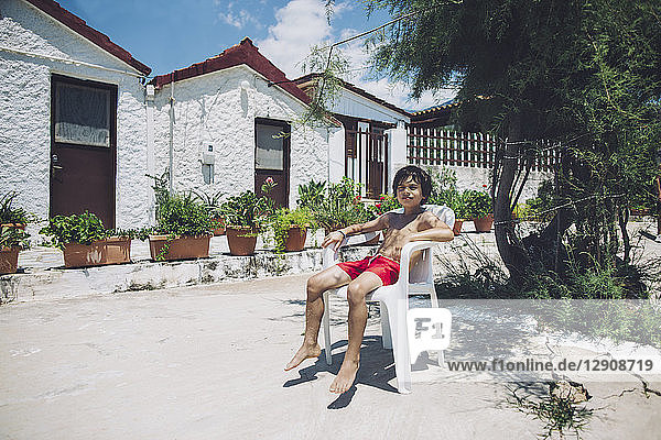 Portrait of a boy sitting in swimming trunks in sunshine in front of a house