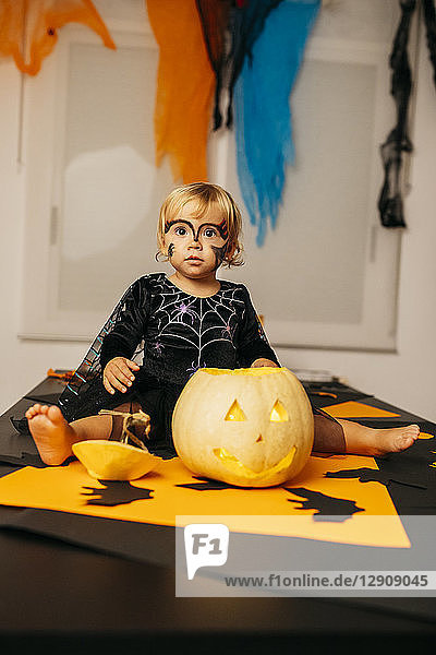 Portrait of little girl with painted face and fancy dress sitting on table with Jack O'Lantern