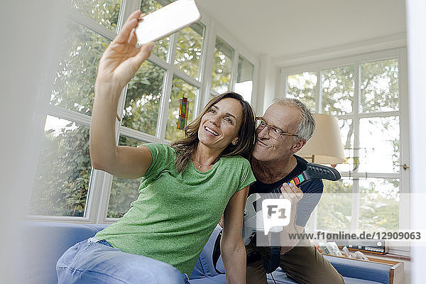 Happy mature couple taking a selfie at home with man playing toy electric guitar