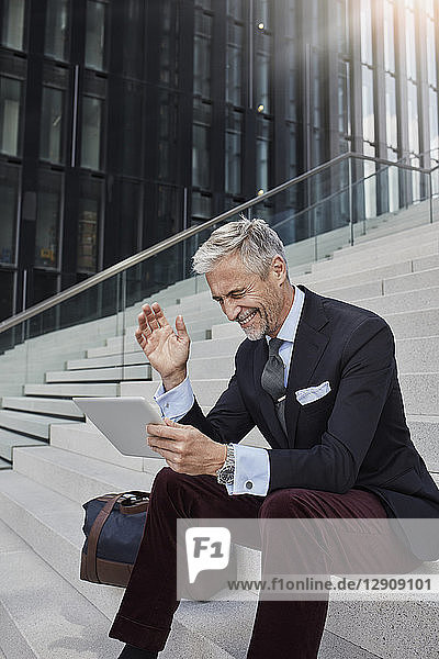 Fashionable businessman with travelling bag sitting on stairs using tablet