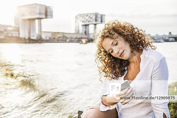 Germany  Cologne  portrait of young woman sitting at riverside looking at cell phone