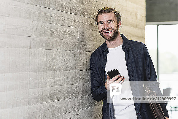 Happy casual businessman standing on office floor holding cell phone and briefcase