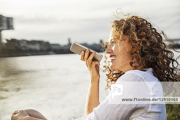 Germany  Cologne  portrait of laughing young woman on the phone sitting at riverside in the evening