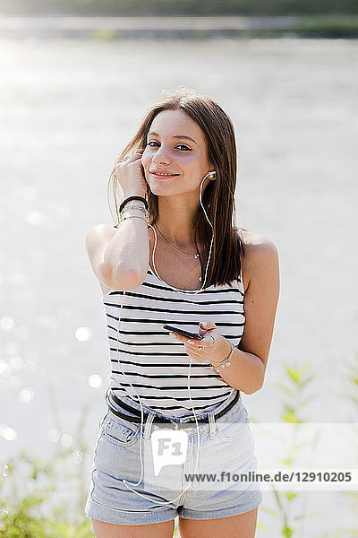 Portrait of smiling young woman listening music with earphones and smartphone at the waterfront