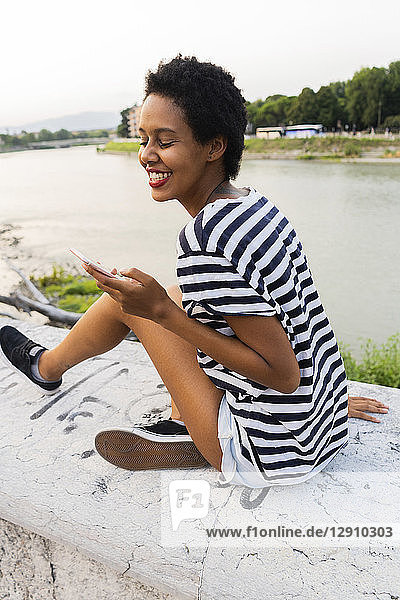Smiling young woman sitting on wall at the riverside using cell phone