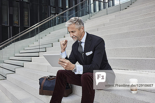 Fashionable businessman with travelling bag nd coffee to go sitting on stairs using tablet